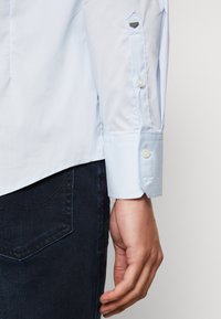 Hellblaues Langarmhemd mit Knopfmanschette, glatter Textur und einfachem Design. Im Hintergrund ist dunkelblauer Denim sichtbar.