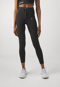 Black high-waisted leggings with a smooth texture, featuring a decorative lace-up front and subtle logo detail on the left thigh.