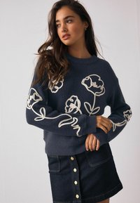 Mørkeblå fleecetrøje med hvid blomsterbroderi på ærmerne. Rund halsudskæring, lange ærmer, afslappet pasform. Kombineret med et denimskørt.