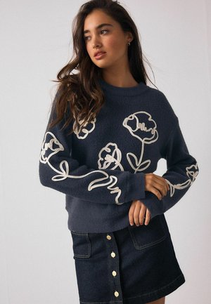 Tumši zils vilnas džemperis ar baltu ziedu izšuvumu uz piedurknēm. Apaļa kakla izgriezuma, garas piedurknes, brīva piegriezuma. Kombinēts ar džinsa svārku.