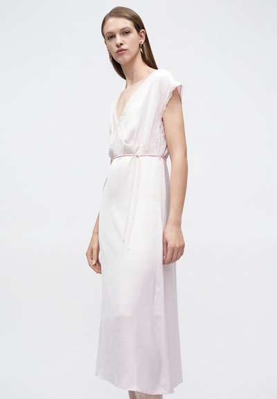 Γυναίκα με ίσια καστανά μαλλιά, ντυμένη με ένα ροζ παστέλ φόρεμα τύπου wrap με δαντέλα στο τελείωμα, δεμένο στη μέση, που στέκεται μπροστά σε απλό λευκό φόντο.