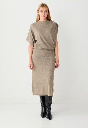 Gebreide jurk in beige met een ontspannen pasvorm, korte dolmanmouwen en een midilange zoom. Gecombineerd met hoge zwarte laarzen, waardoor een slank silhouet ontstaat.