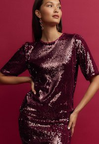 Haut en sequins bourgogne à manches courtes, col rond et texture lisse. Assorti avec une jupe en sequins. Arrière-plan de couleur rose uni.