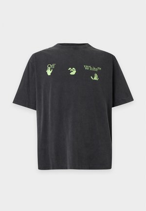 Jeune homme debout face à l'avant portant un t-shirt noir Off-White avec des motifs de mains verts, un pantalon de survêtement beige et des baskets blanches.