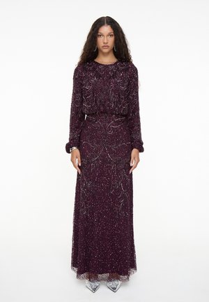 Mujer con pelo largo y rizado, vestida con un vestido de lentejuelas morado oscuro hasta el suelo con mangas largas y zapatos plateados puntiagudos.