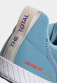 Baskets en daim bleu avec une tige texturée, semelle en caoutchouc blanche et une sangle tissée portant les inscriptions "THE TOTAL" et "ZERO DEGREE DROP" dans des couleurs contrastées.
