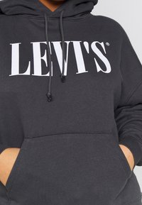 Sweat à capuche Levi's gris foncé avec logo blanc, poche kangourou avant et cordons ajustables, porté avec les mains dans les poches.