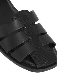 Schwarze Sandale mit flacher Sohle, ausgestattet mit breiten Riemen aus glatten und strukturierten Materialien, offene Zehenpartie und geflochtenen Akzenten.