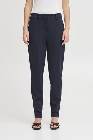 Žena oblečená v priliehavých námornícky modrých nohaviciach a čiernych sandáloch na opätku s remienkami, stojaca pred jednoduchým svetlým pozadím.