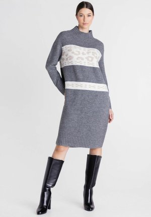 Vrouw die een grijze gebreide sweaterjurk met witte patroonstrepen draagt, gecombineerd met zwarte kniehoge laarzen met hak, staand tegen een effen achtergrond.