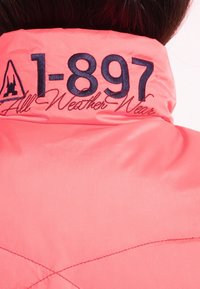 Col de veste corail éclatant avec broderie marine indiquant "1-897" et texte en cursive "All Weather Wear", porté par une personne aux cheveux foncés.