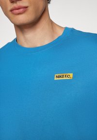Camiseta de algodón azul con cuello redondo, que presenta una etiqueta amarilla "Nike F.C." en el lado izquierdo. Textura suave y corte regular.