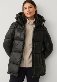 Ung kvinde iført en sort pufferjakke med hætte over en beige zip-up fleece, stående foran en ensfarvet lys væg.