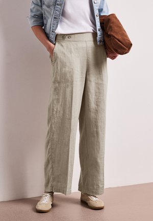 Osoba ubrana w luźne beżowe lniane spodnie, białe trampki, białą koszulkę, jasną jeansową kurtkę, trzymająca złożoną brązową zamszową kurtkę.