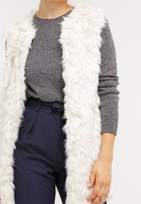 Fuskpälsväst i off-white, med en luddig textur, lång design och öppen front. Kombinerad med en grå tröja och marinblå byxor.