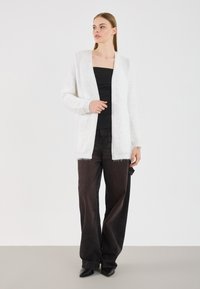 Cardigan alb, pufoasă, cu deschidere în față și mâneci lungi, purtată cu un top negru și pantaloni largi de culoare închisă, pe un fundal neutru.