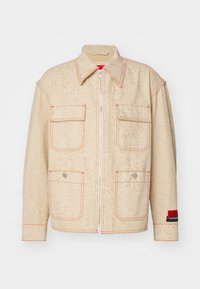 Beige jacka med mönstrad textur, med en framdragkedja, två bröstfickor, röda sömmar och en färgblockerad manschett.