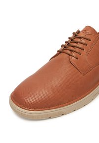 Hnědé kožené boty s perforovanou texturou, kulatou špičkou a šněrováním. Podešev je krémová, což poskytuje kontrast s vrchním materiálem.