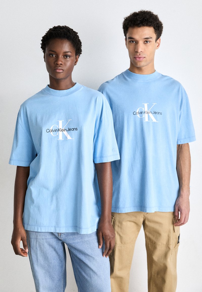Kaksi henkilöä pukeutuu vaaleansinisiin puuvillapaidoihin, joissa on valkoinen "Calvin Klein Jeans" -logo. Toisella on päällään siniset farkut, toisella taas vaaleat cargo-housut.