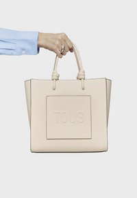 Beżowa skórzana torba tote z wytłoczonym logo "TOUS", o ustrukturyzowanym kształcie, z podwójnymi uchwytami u góry i gładką fakturą. Trzymana w dłoni na jednolitym tle.