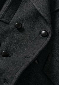 Zwarte wollen jas met een gestructureerde stof, dubbelzijdig ontwerp en ronde zwarte knopen. Close-up van de kraag en sluitingdetails.