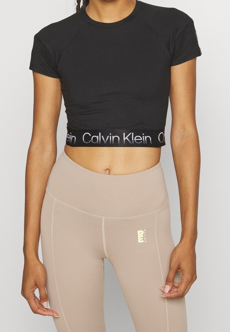 Černý zkrácený sportovní top s krátkými rukávy, se spodním lemem s logem Calvin Klein, kombinovaný s béžovými legínami s vysokým pasem.