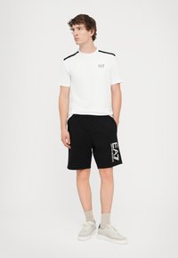 White t-shirt with black shoulder accents and front logo, paired with black shorts featuring a white logo. Worn with light-colored sneakers.