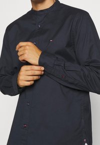 Granatowa koszula z długim rękawem i stójką, z zapinanymi mankietami, subtelnym logo oraz gładkim, teksturowanym materiałem.