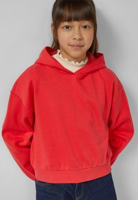 Koralowa bluza z kapturem o luźnym kroju i ściągaczu u dołu. Miękka tkanina z długimi rękawami, noszona na bluzce z wysokim dekoltem. Minimalistyczny design.