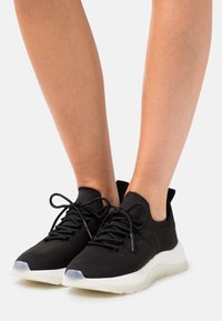 Sneakers in maglia nera con una tomaia leggera e testurizzata, lacci regolari e una suola intermedia bianca. Accento in gomma trasparente sulla punta.