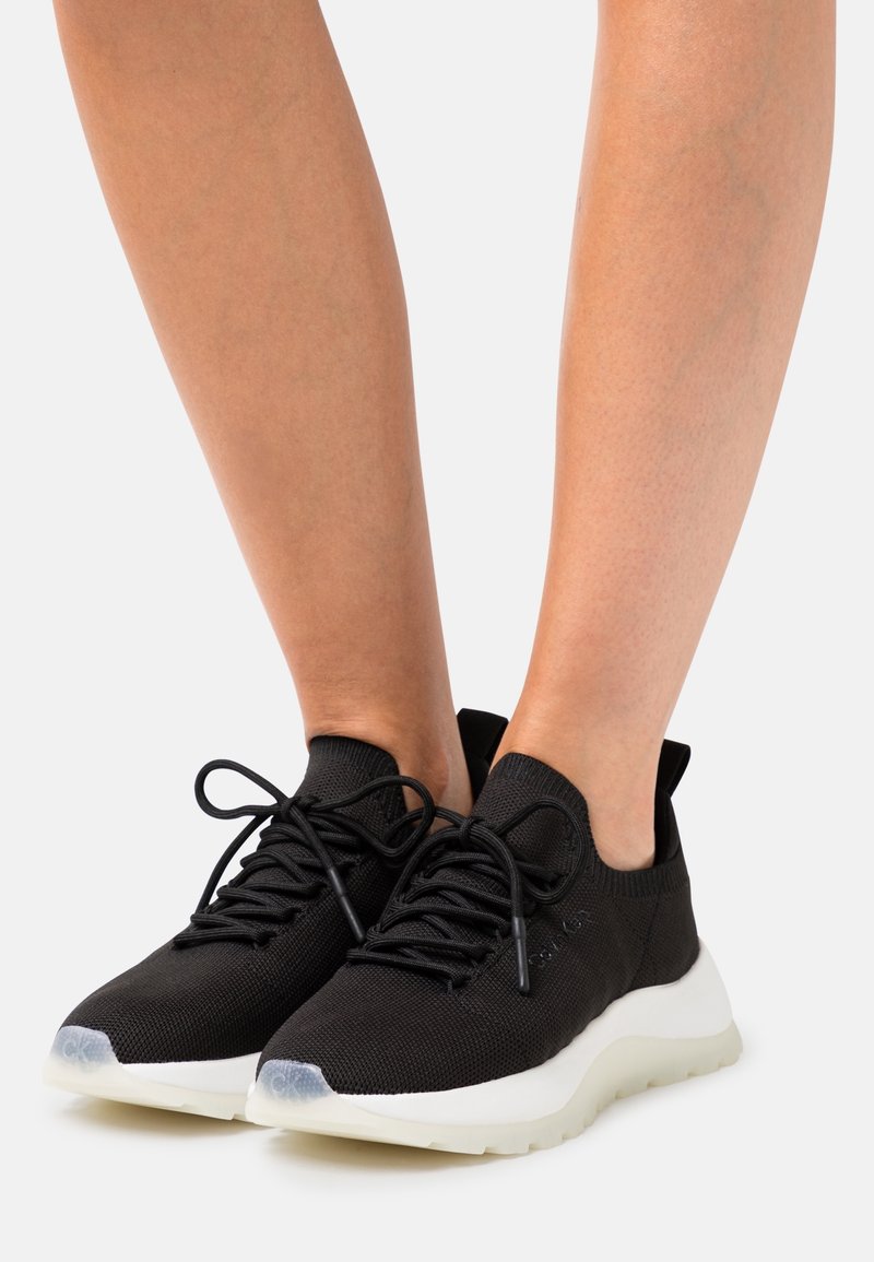 Sneakers in maglia nera con una tomaia leggera e testurizzata, lacci regolari e una suola intermedia bianca. Accento in gomma trasparente sulla punta.