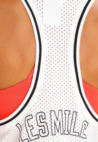 Biały sportowy top bez rękawów z siatkowym wzorem, czarnymi wstawkami i grafiką "LES MILLS" na plecach. Czerwony stanik sportowy widoczny pod spodem.