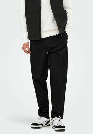 Mann in schwarzen, schmal geschnittenen Hosen, einem weißen gerippten Pullover, einer schwarzen ärmellosen Zip-Weste und weißen sowie schwarzen Sneakers, stehend vor einem weißen Hintergrund.