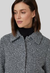 Cappotto grigio testurizzato con collo a giacca e chiusura a bottoni. Il tessuto presenta un motivo riccio ed è sovrapposto a un dolcevita nero.