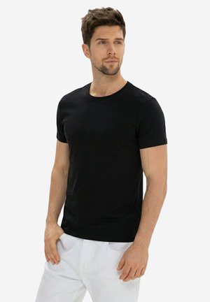 Junger Mann mit kurzen braunen Haaren, der ein schlichtes schwarzes kurzärmeliges Hemd und weiße Hosen trägt, schaut seitlich vor einem weißen Hintergrund.