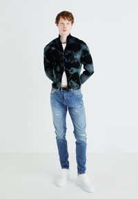 Veste côtelée tie-dye noire et verte avec un col montant, associée à un jean bleu clair déchiré et des baskets blanches. Design minimaliste.