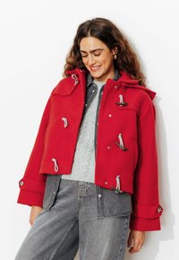 Veste rouge courte avec des fermetures à bascule en argent, une capuche et des détails à cordon. Portée sur un pull en tricot gris et un jean en denim gris clair.