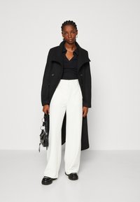 Cappotto lungo nero su una maglietta nera e pantaloni bianchi a gamba larga. Il cappotto ha un colletto alto e spalle arrotondate. Gli accessori includono una piccola borsa nera.