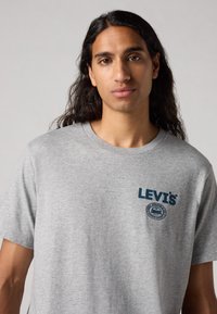 Pilkas medvilnės marškinėliai su apvaliu kaklu, trumpomis rankovėmis ir mėlynu atspaudiniu Levi's logotipu su apvalios formos ženklu ant kairiojo krūtinės.