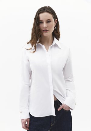 Kobieta o falowanych brązowych włosach, ubrana w białą koszulę zapinaną na guziki i ciemne dżinsy, stoi z jedną ręką w kieszeni na jednolitym białym tle.