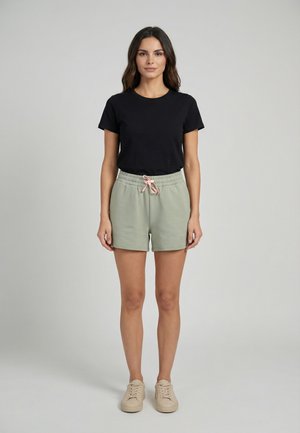 Femme debout, de face, portant un t-shirt noir, un short vert clair avec un cordon de serrage rose et des baskets beiges sur un fond uni.