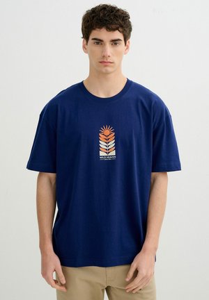 Joven con camiseta azul marino con diseño de planta en naranja y blanco y texto "WILD HEAVEN", combinado con pantalones beige.