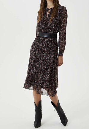 Nainen pukeutuneena mustaan kukalliseen midi-mekkoon, jossa on läpikuultavat pitkät hihat, leveä musta vyö ja mustat terävät nilkkurit.