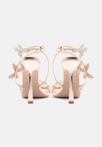 Sandały na wysokim obcasie w kolorze różowego złota z połyskującym wykończeniem, motylkowymi ozdobami i cienkimi paskami wokół kostki. Posiadają szpilkowy obcas i miękką teksturę.