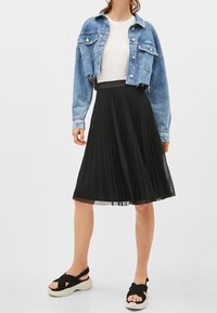 Kvinna som bär en vit topp, en kort blå denimjacka, en svart plisserad midikjol och svarta plattformssandaler med tjocka remmar.