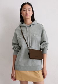 Szary hoodie z haftowanym tekstem, w zestawie z beżową spódnicą. Na ramieniu wisi mała brązowa torebka na ramię z paskiem.