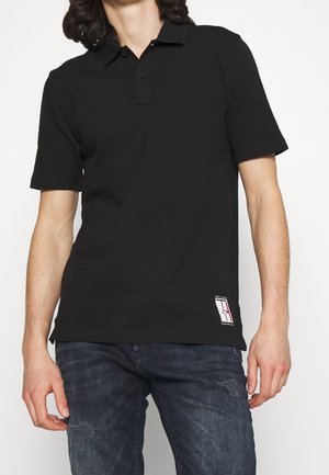 Sort polo-shirt lavet af bomuld med korte ærmer, en tre-knaps placket, og et lille logo-label nær kanten. Includeret med slidte blå jeans.