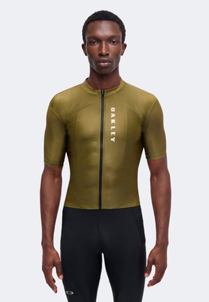 Hombre con un maillot de ciclismo de manga corta de color verde oliva de Oakley con cremallera frontal y pantalones cortos de ciclismo negros.