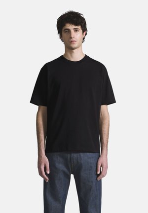 Jeune homme debout portant un t-shirt noir uni à manches courtes et un jean bleu sur un fond gris clair.