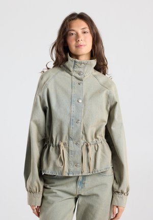 Jonge vrouw met lang haar draagt een lichtgrijze jas met knopen, hoge kraag en trekkoorden, gecombineerd met bijpassende broek.